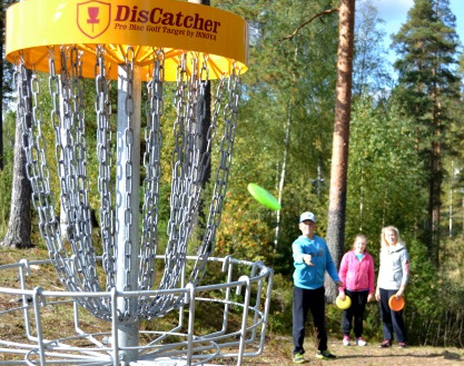 Kuvassa frisbeegolf kori ja kolme frisbeen heittäjää.